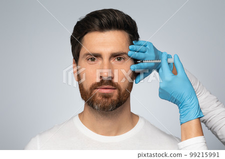 Handsome bearded man getting under eye injections in beauty clinic, looking at camera, standing on grey background Handsome bearded man getting under eye injections in beauty clinic, looking at camera, standing on grey background 99215991