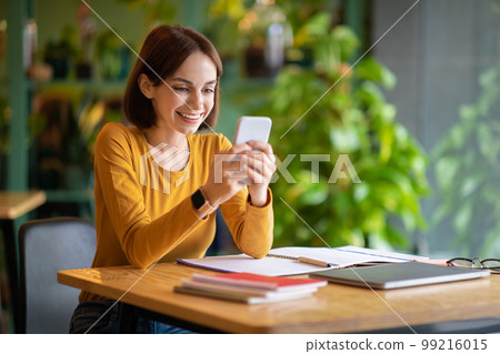 Cheerful attractive young brunette woman using phone at cafe 99216015