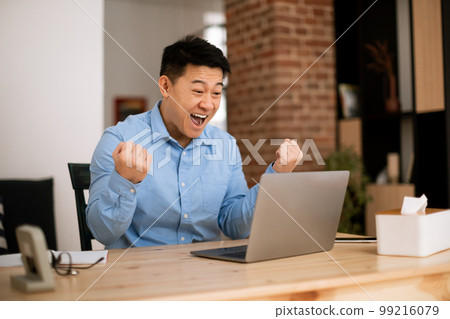 Portrait of excited asian middle aged man using laptop and shaking clenched fists, sitting at desk at home office 99216079