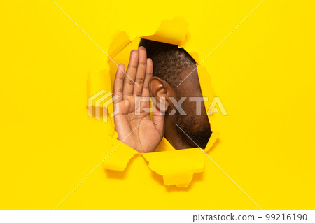 Eavesdropping. Unrecognizable black guy putting ear to torn yellow paper background, listening through hole, closeup 99216190