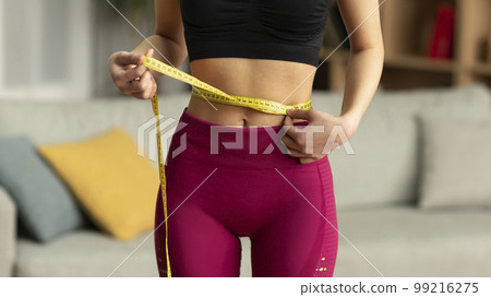 Weight loss concept. Cropped view of fit lady measuring her waist using yellow tape, checking her body parameters 99216275
