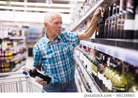 old age man examines bottle of vermouth in alcoholic section of supermarket old age man examines bottle of vermouth in alcoholic section of supermarket 99216665