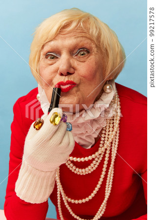Applying lipstick. Beautiful old woman, grandmother in stylish red dress and pearl necklace posing over blue studio background. Concept of age, fashion and emotions 99217578