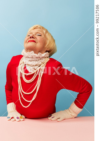 Delightful. Beautiful old woman, grandmother in stylish red dress and pearl necklace posing over blue studio background. Concept of age, fashion and emotions 99217586