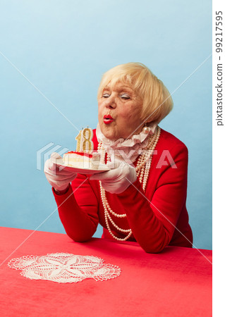 Making wish. Beautiful old woman, grandmother in stylish red dress and pearl necklace posing with birthday cake over blue studio background. Concept of age, fashion and emotions 99217595