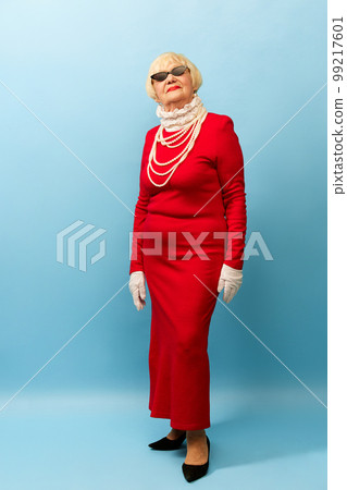 Fashionable look. Beautiful old woman, grandmother in stylish red dress and pearl necklace posing over blue studio background. Concept of age, fashion and emotions 99217601