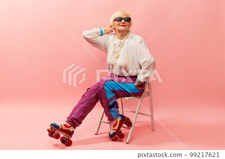 Retro style. Beautiful old woman, grandmother in stylish sportive trousers posing on vintage rollers over pink studio background. Concept of age, fashion and emotions 99217621