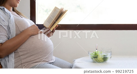 Young pregnant woman sitting reading a book and eating salad at home Young pregnant woman sitting reading a book and eating salad at home 99219725
