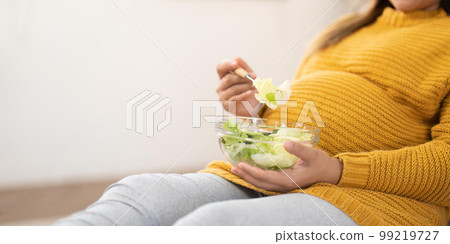 Happy pregnant young woman sitting and eating salad at home Happy pregnant young woman sitting and eating salad at home 99219727