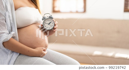 An Asian pregnant woman in comfortable clothes sits on a chair in her living room, holding her belly and alarm clock. awaiting baby, baby's due date concept 99219742