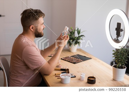 Portrait of happy man beauty blogger sitting in room at table and speaking recommending foundation or decorative cosmetic looking at camera. Blogging and vlog concept 99220338