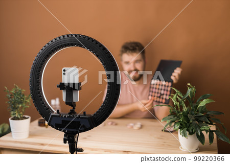 Portrait of happy man beauty blogger sitting in room at table and speaking recommending foundation or decorative cosmetic looking at camera. Blogging and vlog concept 99220366
