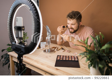 Famous blogger. Cheerful male vlogger showing cosmetics products while recording video and giving advices for his beauty blog. Make-up artist and recording beauty vlog concept Famous blogger. Cheerful male vlogger showing cosmetics products while recording video and giving advices for his beauty blog. Make-up artist and recording beauty vlog concept 99220661