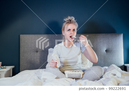 Shocked, stressed, surprised woman eating ice cream, holding a remote control while watching TV in bed. Scary horror movie or terrible news, scare in her eyes Overeating, Jamming of negative emotions Shocked, stressed, surprised woman eating ice cream, holding a remote control while watching TV in bed. Scary horror movie or terrible news, scare in her eyes Overeating, Jamming of negative emotions 99222691