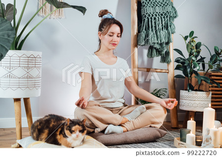Young healthy woman practicing meditation at home, sitting in lotus position on floor cushion with relaxed cat pet in modern Scandi interior home. Meditating, relaxed, eyes closed. Selective focus. 99222720