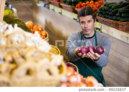 Male seller holding red onion standing in fruit and vegetable section of supermarket 99228837