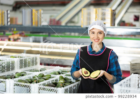 Smiling female worker in fruit sorting warehouse holding sliced avocado 99228934