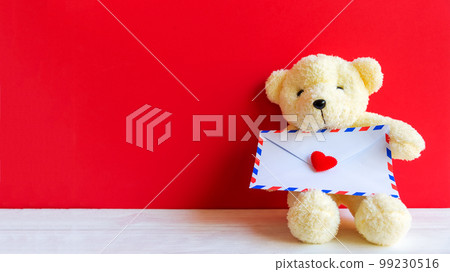 Cute teddy bear clutching love letter in its arms on wooden red background, copy space. Cute teddy bear clutching love letter in its arms on wooden red background, copy space. 99230516