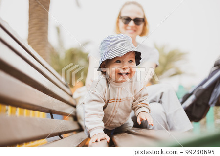 Young mother with her cute infant baby boy child on bench on urban children's playground on warm summer day Young mother with her cute infant baby boy child on bench on urban children's playground on warm summer day 99230695