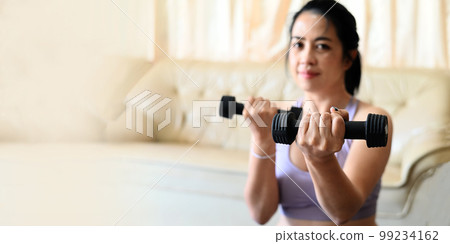 sporty woman exercise muscles in sportswear with dumbbell at home in the living room, woman exercising at home, woman dumbbells in her hands, sport recreation health care concept 99234162
