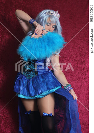 Woman With Blue Hair and Blue Corset Dress Holding Blue Fan on Red Velvet Background Woman With Blue Hair and Blue Corset Dress Holding Blue Fan on Red Velvet Background 99235502