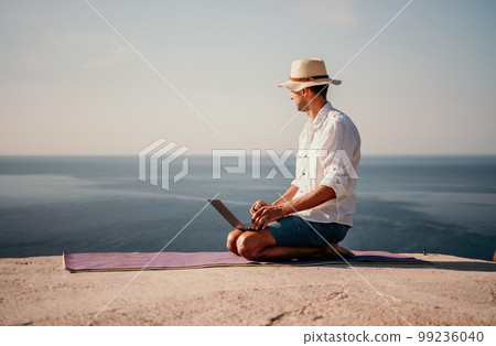 A digital nomad, a man in a hat, a businessman with a laptop does yoga on the rocks by the sea at sunset time, does a business operation online from a distance. Remote work on vacation. 99236040