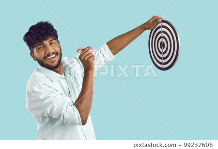 Portrait of smiling Indian guy aiming dart at dart board on light blue background. 99237608