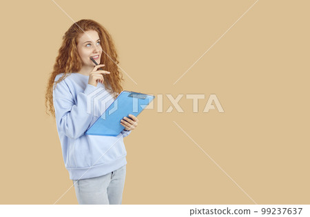 Woman standing on beige copy space background, holding clipboard, biting pen and thinking 99237637