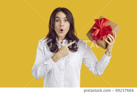 Surprised amazed brunette woman pointin on Christmas gift in her hand on yellow background. Surprised amazed brunette woman pointin on Christmas gift in her hand on yellow background. 99237668
