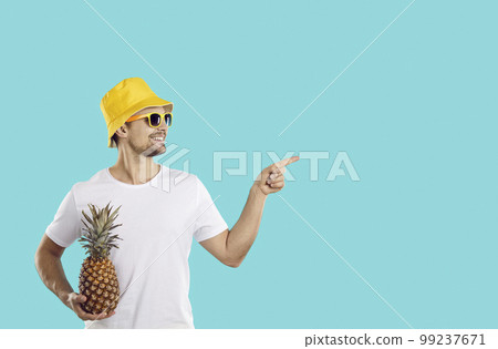 Cheerful young Caucasian man in sunglasses holding pineapple and pointing with finger at copy space Cheerful young Caucasian man in sunglasses holding pineapple and pointing with finger at copy space 99237671