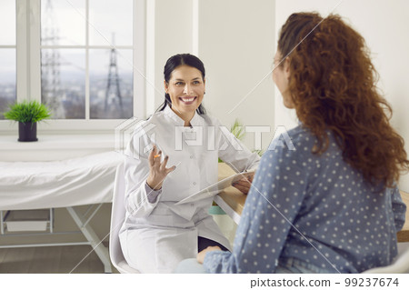 Happy doctor with clipboard talking to young woman during medical consultation at clinic Happy doctor with clipboard talking to young woman during medical consultation at clinic 99237674