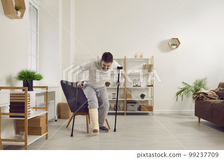 Man with a broken leg tries to stand up off his chair and walk with a stick and crutches Man with a broken leg tries to stand up off his chair and walk with a stick and crutches 99237709