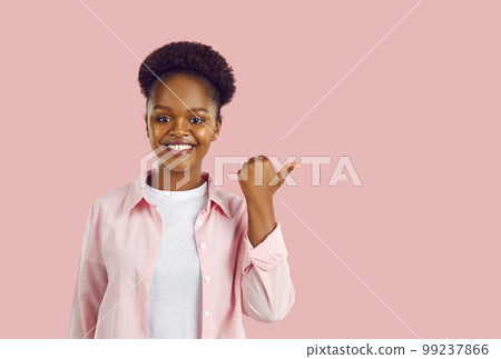 A happy young African-American woman with a smile points her thumb to the side, pink background. 99237866