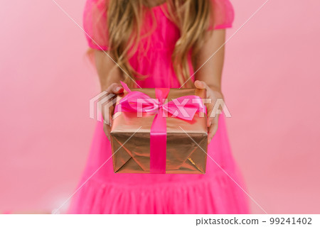 Close up of a girl holding a gift box with a pink silk ribbon, giving a gift for a holiday. Merry Christmas, Happy New Year, Valentine's Day, holiday, festive, Birthday, surprise concept. 99241402