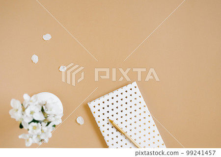 A white notebook with a gold pen on a beige table and flowers in a defocus vase. Blogger's workplace. Top view, copy space 99241457