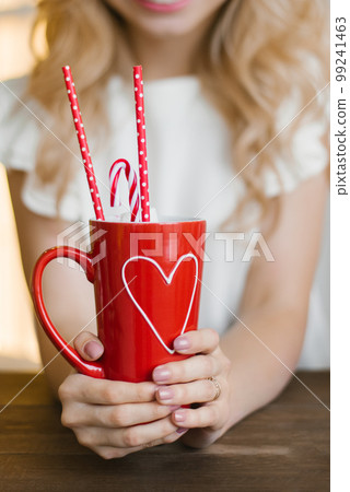 A red mug with a heart pattern with cocoa and paper tubes in the hands of a woman. Valentine's Day Concept 99241463