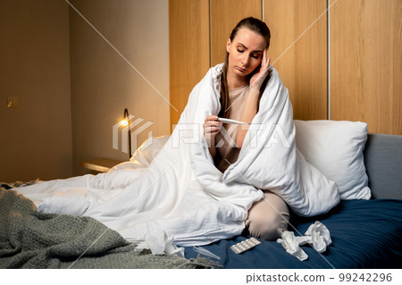 A sick, flu-ridden young woman, a girl has a headache, she has a fever, flu and check the thermometer, measure body temperature, feel the disease while sitting on the bed A sick, flu-ridden young woman, a girl has a headache, she has a fever, flu and check the thermometer, measure body temperature, feel the disease while sitting on the bed 99242296