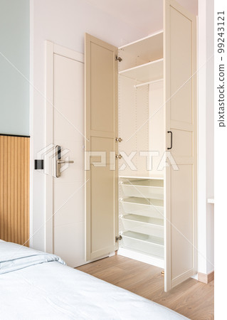 Built-in wooden wardrobe with open doors in beige. Inside the cabinet, all shelves, drawers, crossbars are made of white durable plastic. Entrance to the hotel room with an electronic lock. 99243121