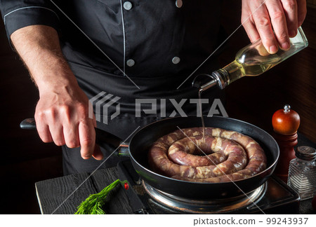 The chef adds oil to pan with raw meat sausage. Preparation for cooking sausages in the kitchen of a restaurant or cafe on table with vegetables 99243937