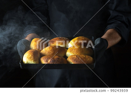 The chef holds a baking sheet with freshly baked buns or pies. Black space for menu or recipe. 99243939
