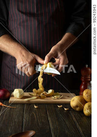 The cook cuts raw potatoes into pieces with a knife before preparing breakfast or dinner. Close-up of a chef hands while working in a kitchen. The cook cuts raw potatoes into pieces with a knife before preparing breakfast or dinner. Close-up of a chef hands while working in a kitchen. 99243943