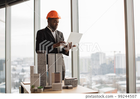Chief architect african man in business suit and protective helmet working with city buildings mode Chief architect african man in business suit and protective helmet working with city buildings mode 99244507