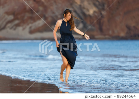 Young woman in long dark blue dress walking through waves on the beach 99244564