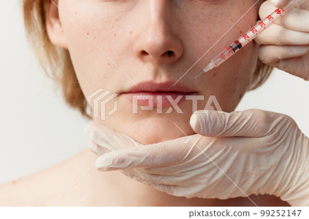 Young redhead man having cosmetological injections. Male model posing against grey background. Lip augmentation 99252147