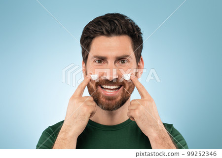 Closeup portrait of handsome bearded man applying moisturising cream on cheeks, standing over blue studio background Closeup portrait of handsome bearded man applying moisturising cream on cheeks, standing over blue studio background 99252346