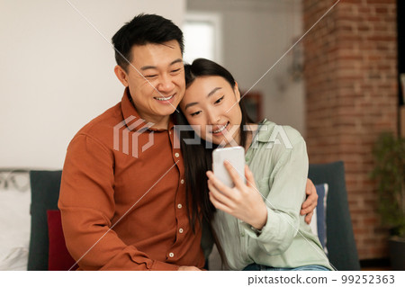 Loving happy korean spouses video calling via cellphone, middle aged man and his young wife relaxing on sofa at home 99252363