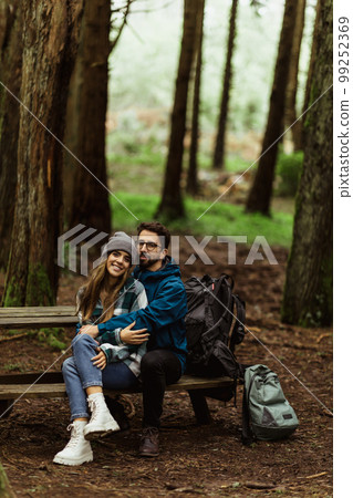 Glad millennial caucasian husband in jacket hugging wife, resting on bench in cold forest with backpack 99252369