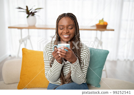 Joyful young black woman sitting on couch, drinking tea 99252541