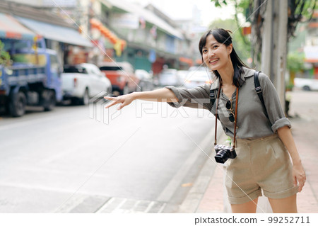 Smiling young Asian woman traveler hitchhiking on a road in the city. Life is a journey concept. 99252711