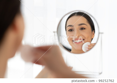 Young Smiling Indian Lady Cleansing Skin With Cotton Pad Near Mirror 99252762